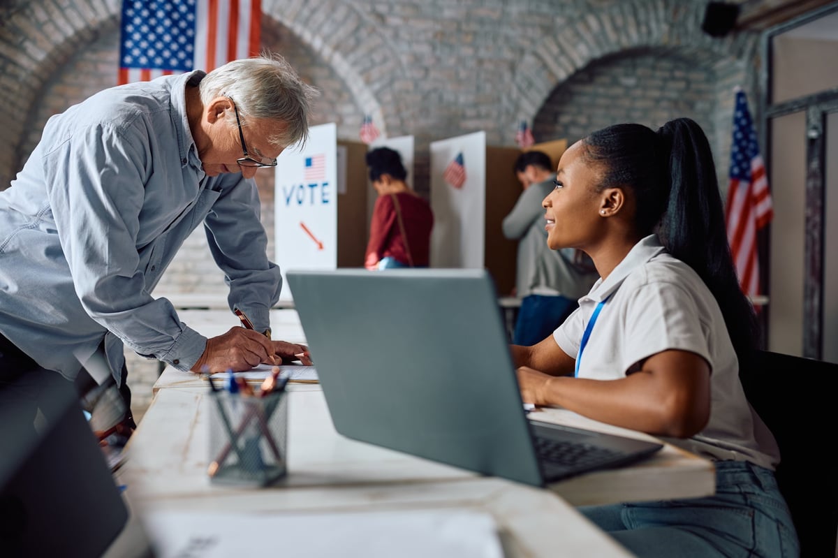 African American polling place volunteer assisting senior man to sign up for voting for the elections.