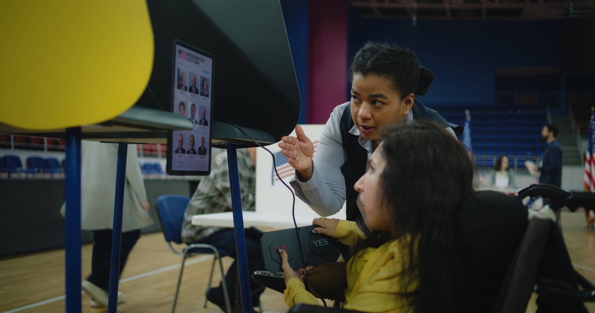 Polling officer consults woman with physical disability. Female US voter with SMA uses remote buttons and votes on tablet computer in voting booth at polling station. Election Day in United States.
