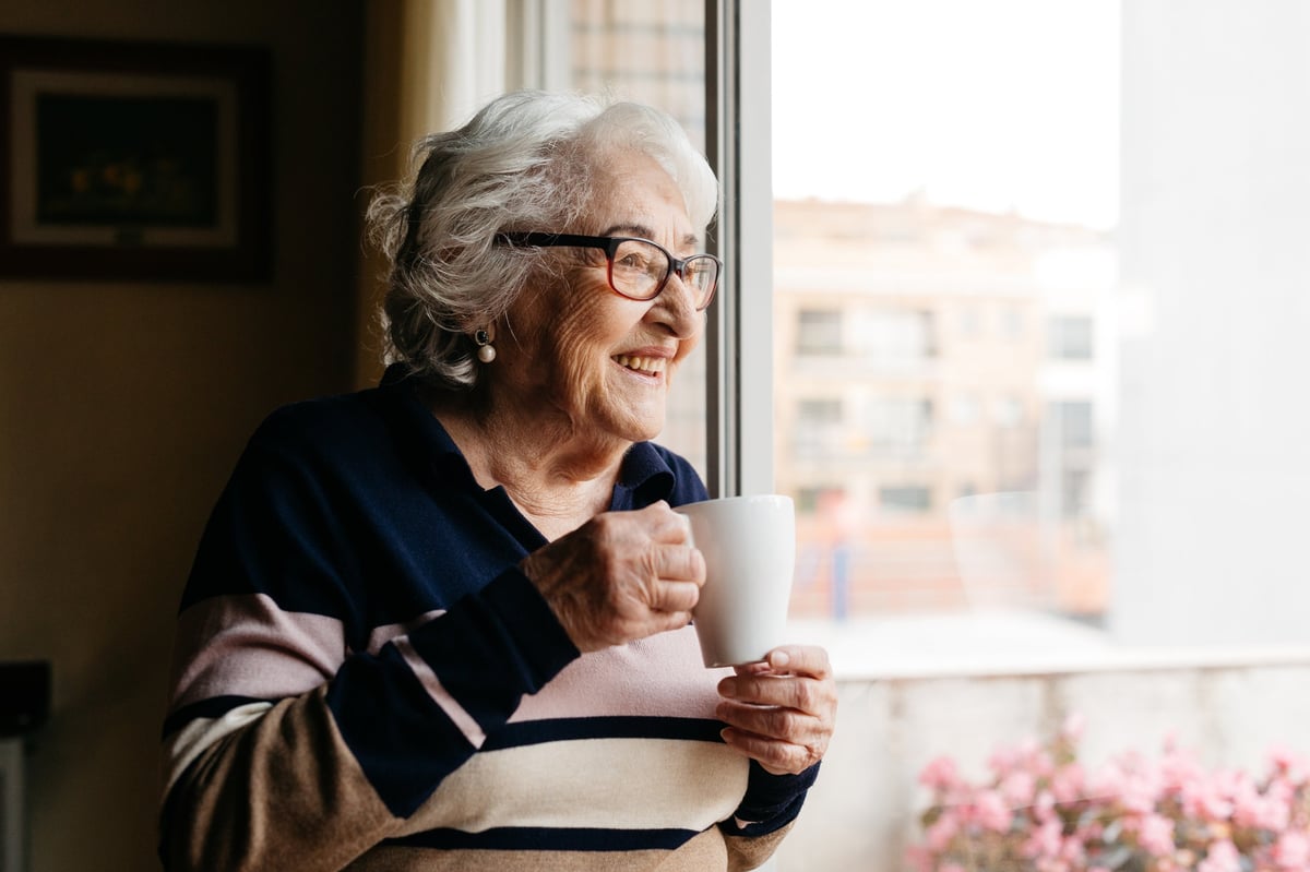 Portrait of an elderly woman holding a cup of coffee and smiling warmly while looking out a window