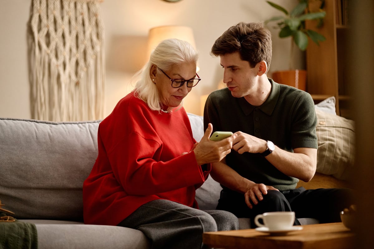 Senior Caucasian woman using smartphone while young adult man guiding her on sofa, showing digital support and tech learning