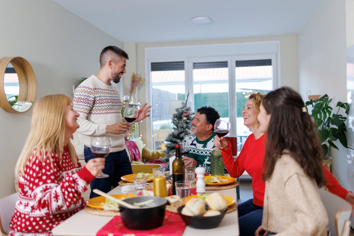 Group of multi-generational family members gathering around a dining table, laughing and making a toast with red wine glasses during a holiday celebration at home