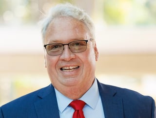 Smiling older man with white hair and glasses wearing a dark blue suit and red tie